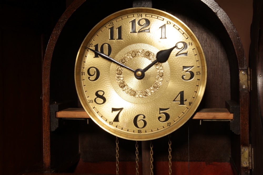A dark oak longcase clock featuring carved arch cresting, a brass dial with Arabic numerals, and a glass trunk door displaying brass weights and pendulum. View VII
