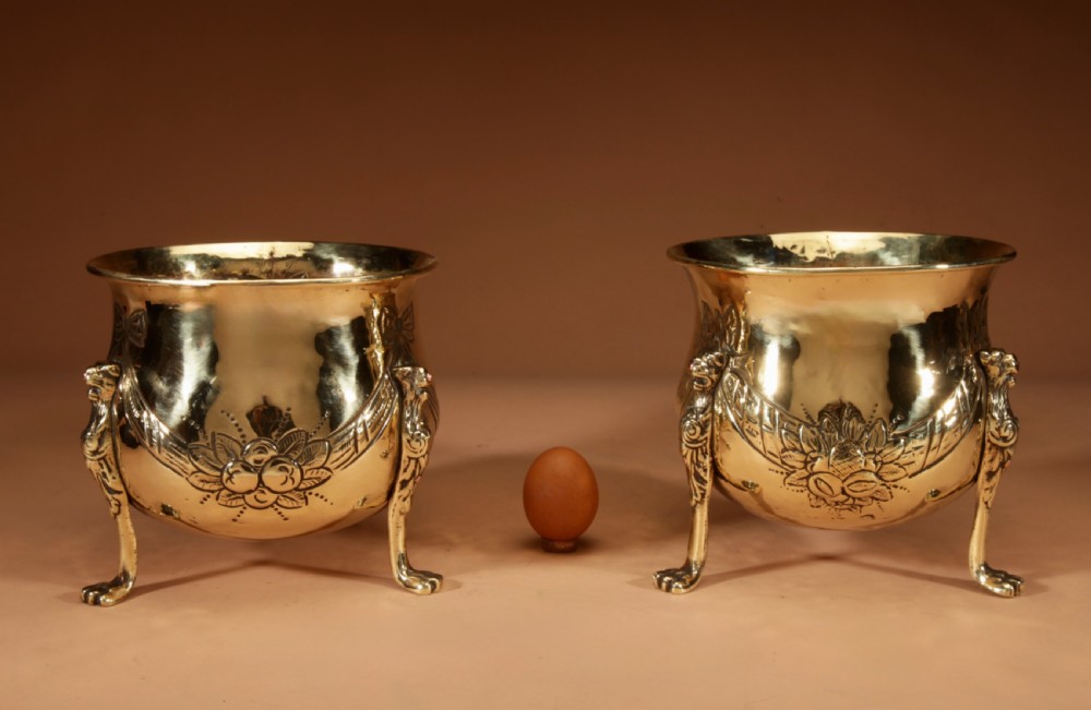A pair of polished brass jardinieres featuring embossed floral swags and resting on tripod lion paw feet.
