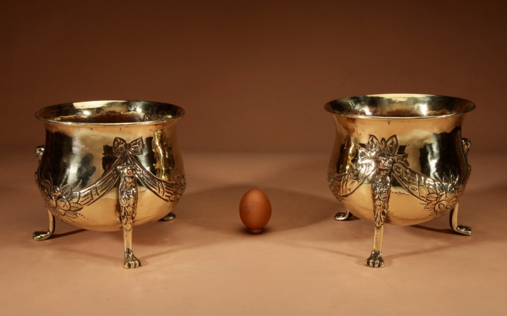A pair of polished brass jardinieres featuring embossed floral swags and resting on tripod lion paw feet. View II