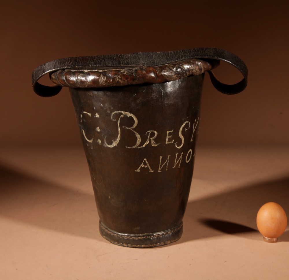 An 18th-century dark brown leather fire bucket featuring a tall stitched leather handle, a thick rolled rim, and hand-painted cream lettering reading BRESYL ANNO 1776. View IV