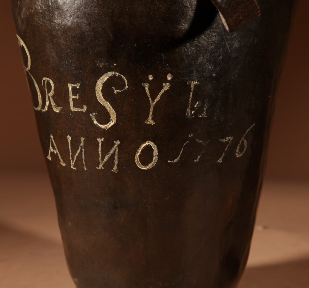 An 18th-century dark brown leather fire bucket featuring a tall stitched leather handle, a thick rolled rim, and hand-painted cream lettering reading BRESYL ANNO 1776. View V