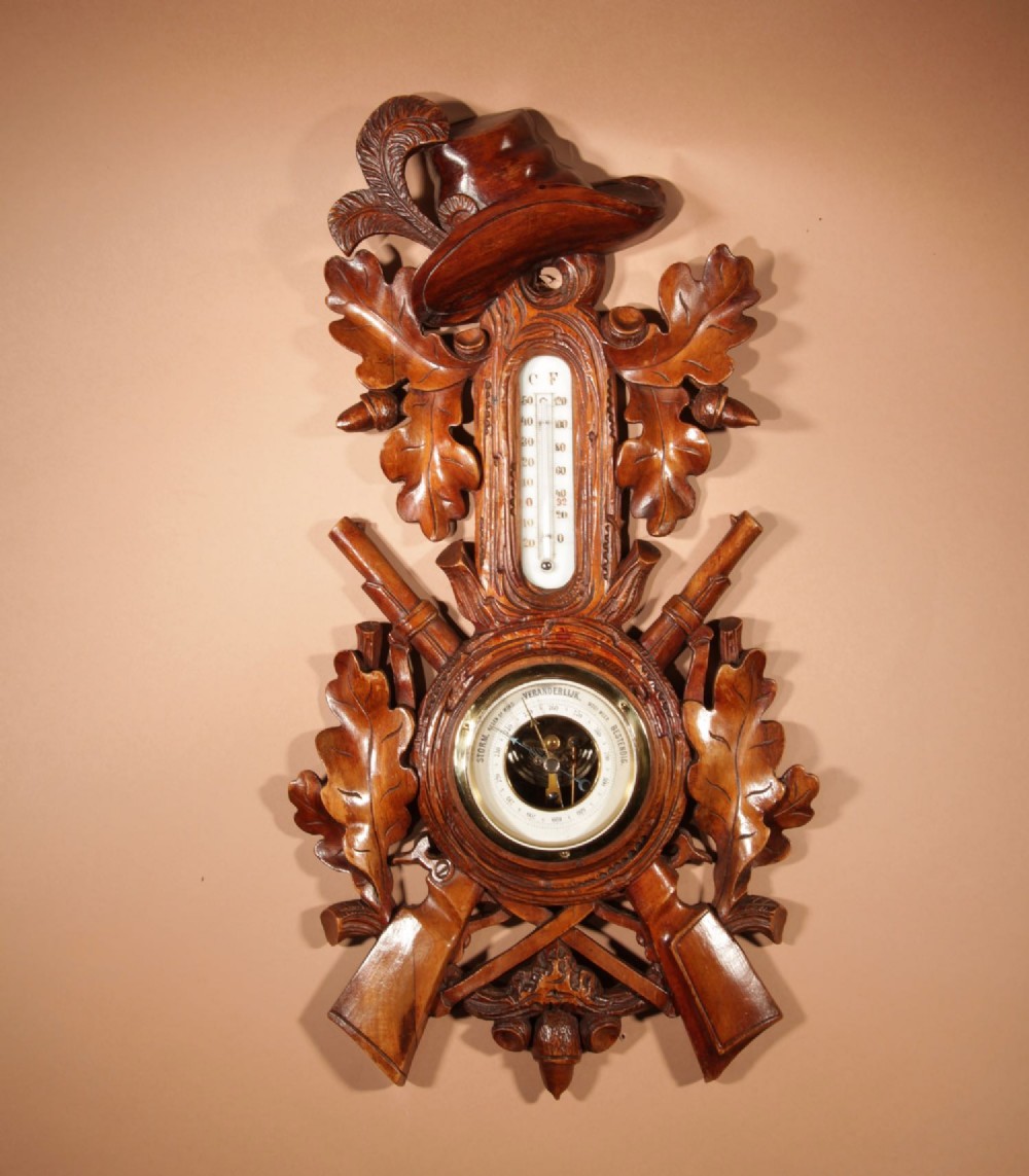 A carved walnut wall-hanging aneroid barometer with a thermometer scale, featuring oak leaves, acorns, crossed rifles, and a carved hat top motif.