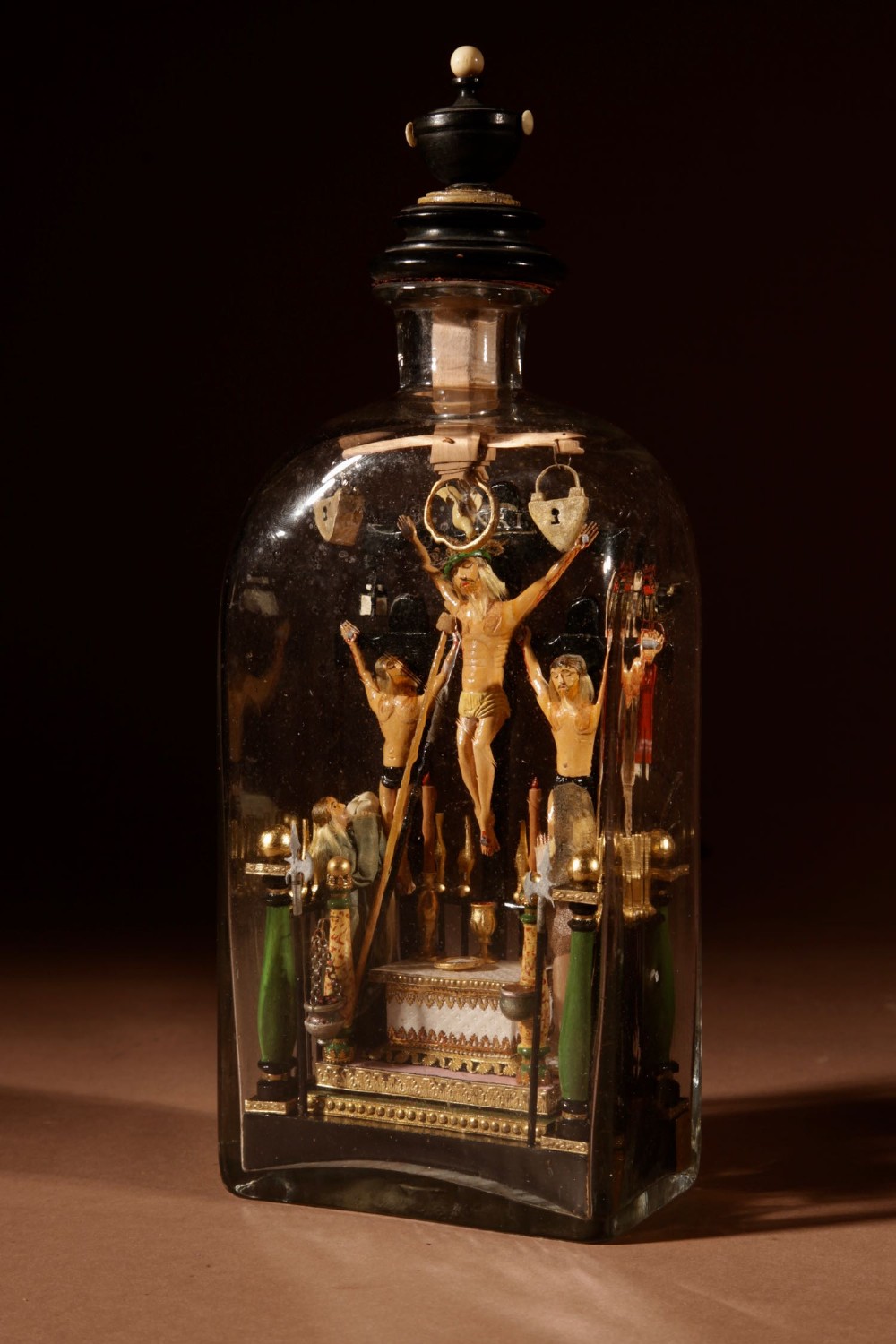 An antique clear glass bottle housing an intricate hand-carved polychrome wooden scene depicting the Crucifixion flanked by figures and religious instruments.