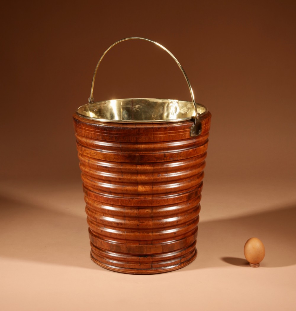 A tapered, coopered walnut bucket featuring horizontal ribbed bands, a fitted brass liner, and a brass swing handle.