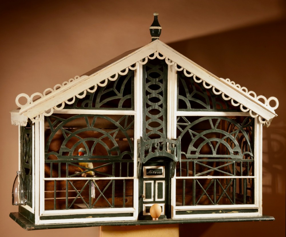 An Edwardian architectural wooden birdcage featuring dark green fretwork panels, cream trim, a gabled roof, and a miniature balcony stamped with the year 1914.