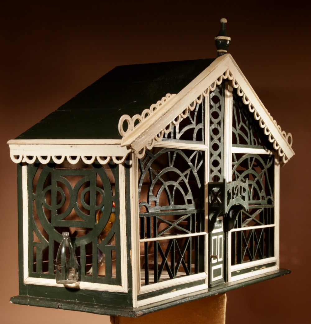 An Edwardian architectural wooden birdcage featuring dark green fretwork panels, cream trim, a gabled roof, and a miniature balcony stamped with the year 1914. View III