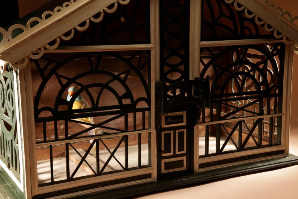 An Edwardian architectural wooden birdcage featuring dark green fretwork panels, cream trim, a gabled roof, and a miniature balcony stamped with the year 1914. View XII