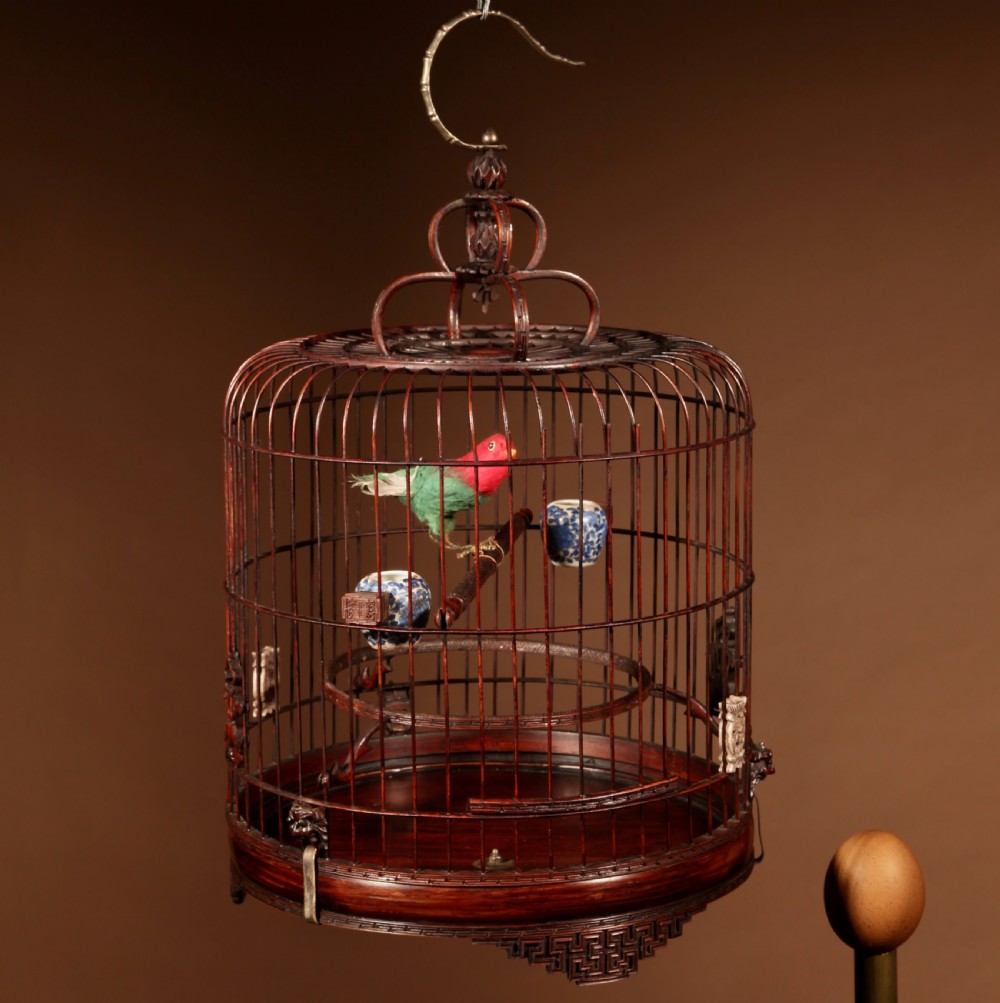 A cylindrical dark brown bamboo birdcage featuring a metal hanging hook, carved base trim, internal perch, and miniature blue and white porcelain cups.