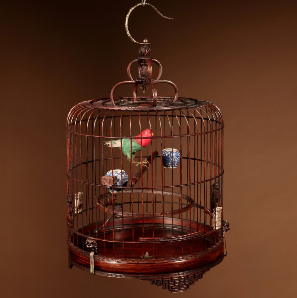 A cylindrical dark brown bamboo birdcage featuring a metal hanging hook, carved base trim, internal perch, and miniature blue and white porcelain cups. View II
