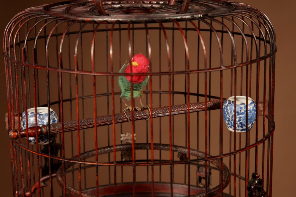 A cylindrical dark brown bamboo birdcage featuring a metal hanging hook, carved base trim, internal perch, and miniature blue and white porcelain cups. View III