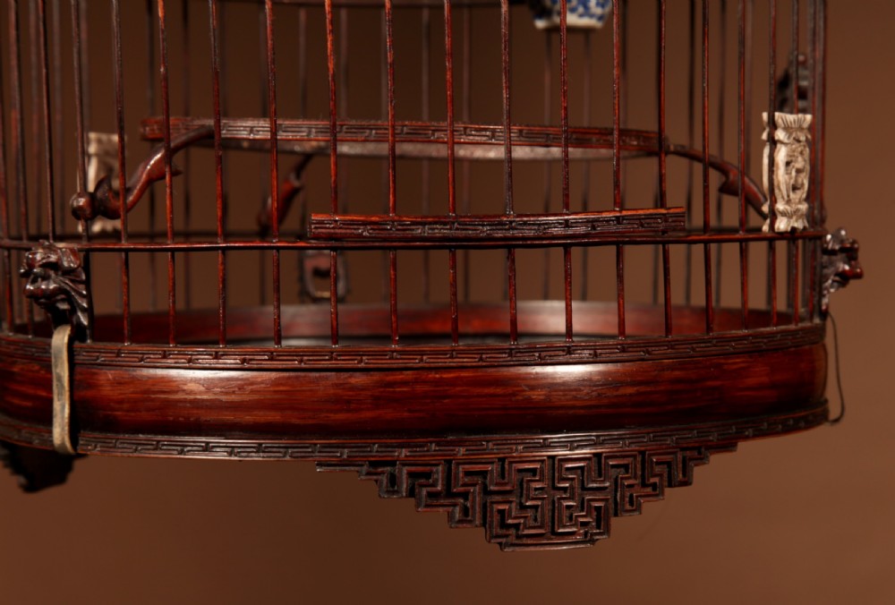 A cylindrical dark brown bamboo birdcage featuring a metal hanging hook, carved base trim, internal perch, and miniature blue and white porcelain cups. View IV