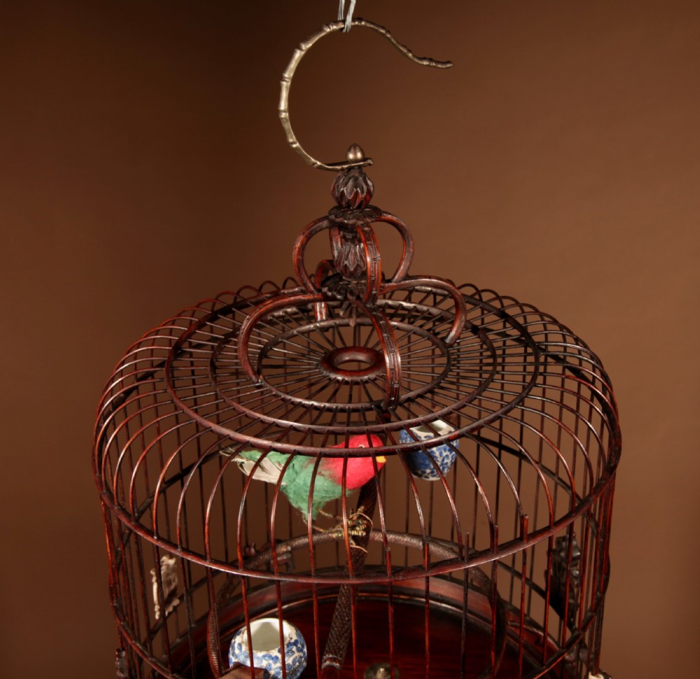 A cylindrical dark brown bamboo birdcage featuring a metal hanging hook, carved base trim, internal perch, and miniature blue and white porcelain cups. View V