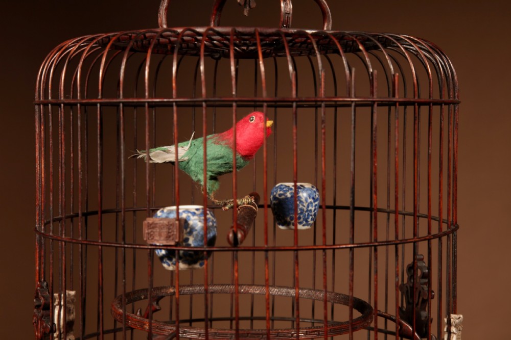 A cylindrical dark brown bamboo birdcage featuring a metal hanging hook, carved base trim, internal perch, and miniature blue and white porcelain cups. View VI