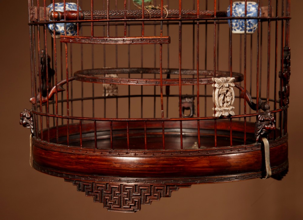 A cylindrical dark brown bamboo birdcage featuring a metal hanging hook, carved base trim, internal perch, and miniature blue and white porcelain cups. View VIII