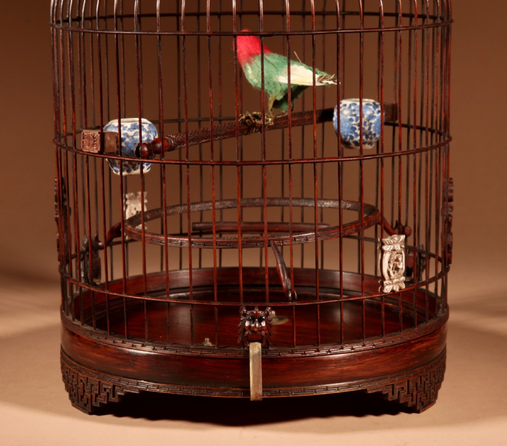 A cylindrical dark brown bamboo birdcage featuring a metal hanging hook, carved base trim, internal perch, and miniature blue and white porcelain cups. View XII