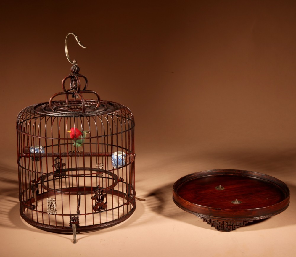 A cylindrical dark brown bamboo birdcage featuring a metal hanging hook, carved base trim, internal perch, and miniature blue and white porcelain cups. View XIV
