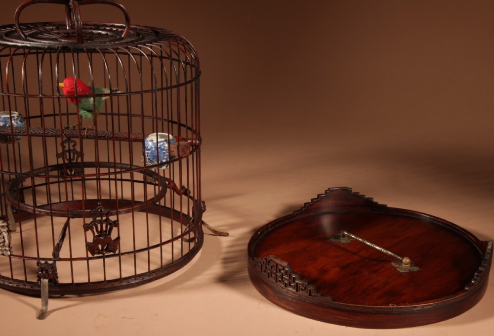 A cylindrical dark brown bamboo birdcage featuring a metal hanging hook, carved base trim, internal perch, and miniature blue and white porcelain cups. View XV