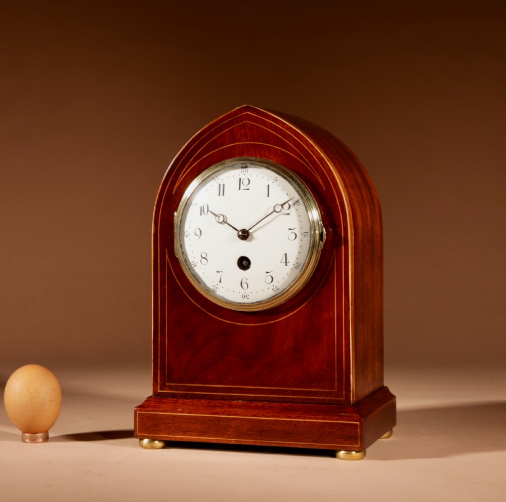 An Edwardian inlaid mahogany mantel clock with a lancet arch top, white enamel dial with Arabic numerals, and brass bun feet.