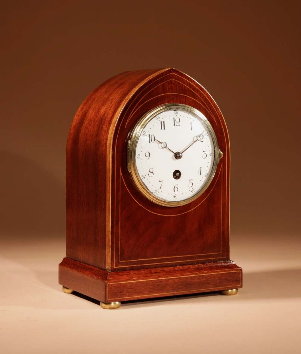 An Edwardian inlaid mahogany mantel clock with a lancet arch top, white enamel dial with Arabic numerals, and brass bun feet. View VI