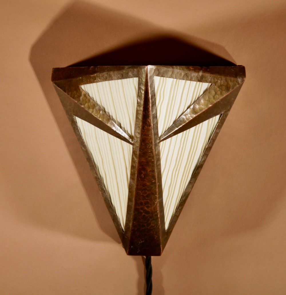An Amsterdam School style triangular copper wall sconce featuring hand-chased geometric metalwork and pleated cream fabric panels.