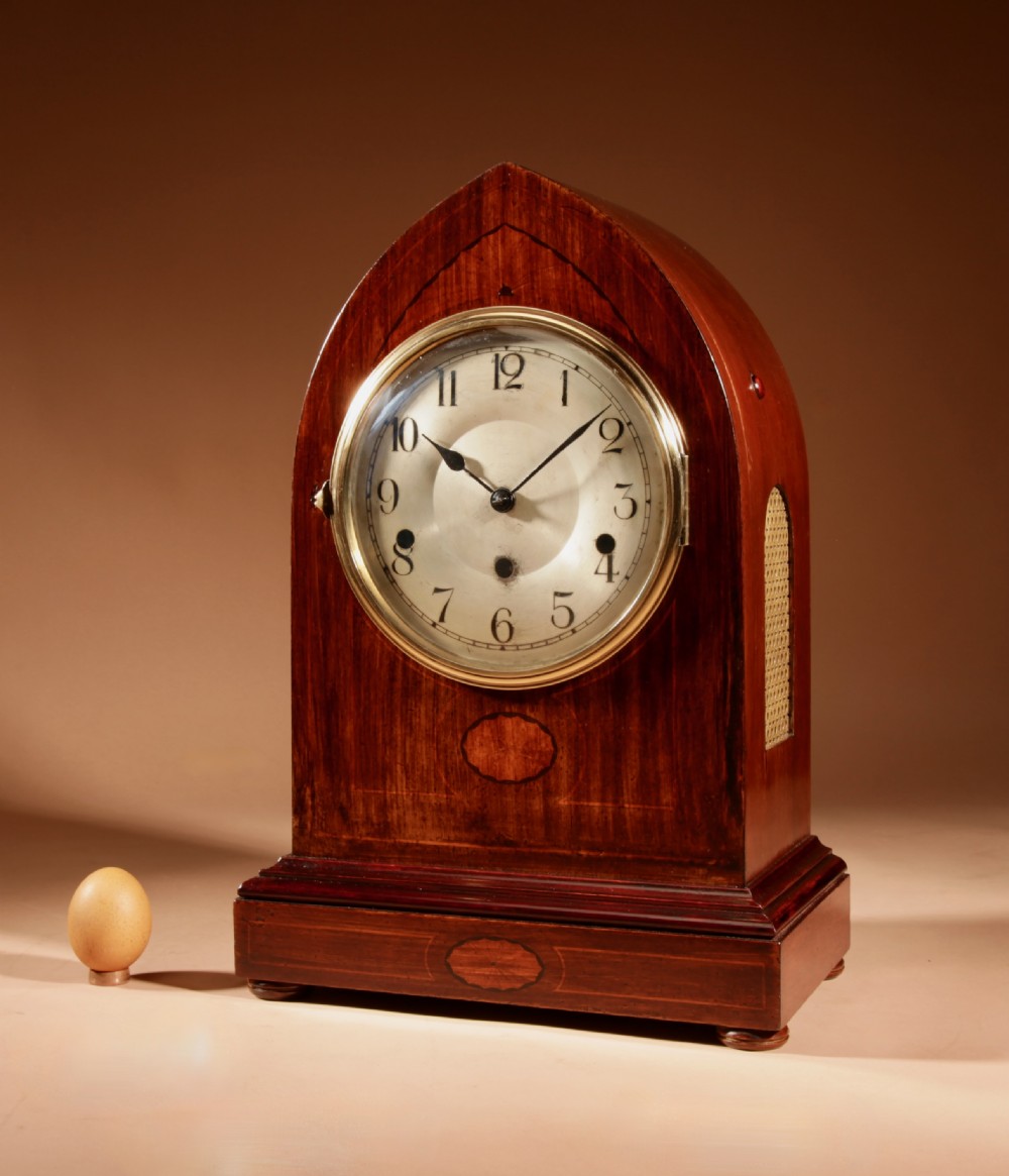 An Edwardian mahogany mantel clock featuring a pointed lancet arch top, oval marquetry inlay, and a circular white dial framed in brass.