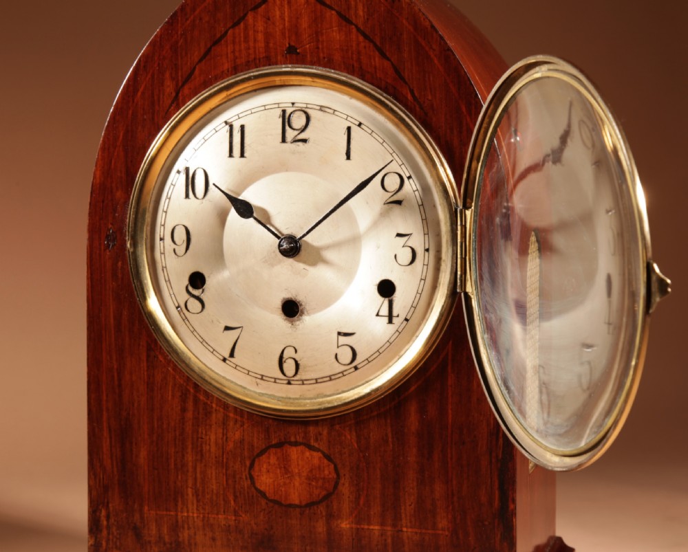 An Edwardian mahogany mantel clock featuring a pointed lancet arch top, oval marquetry inlay, and a circular white dial framed in brass. View IV