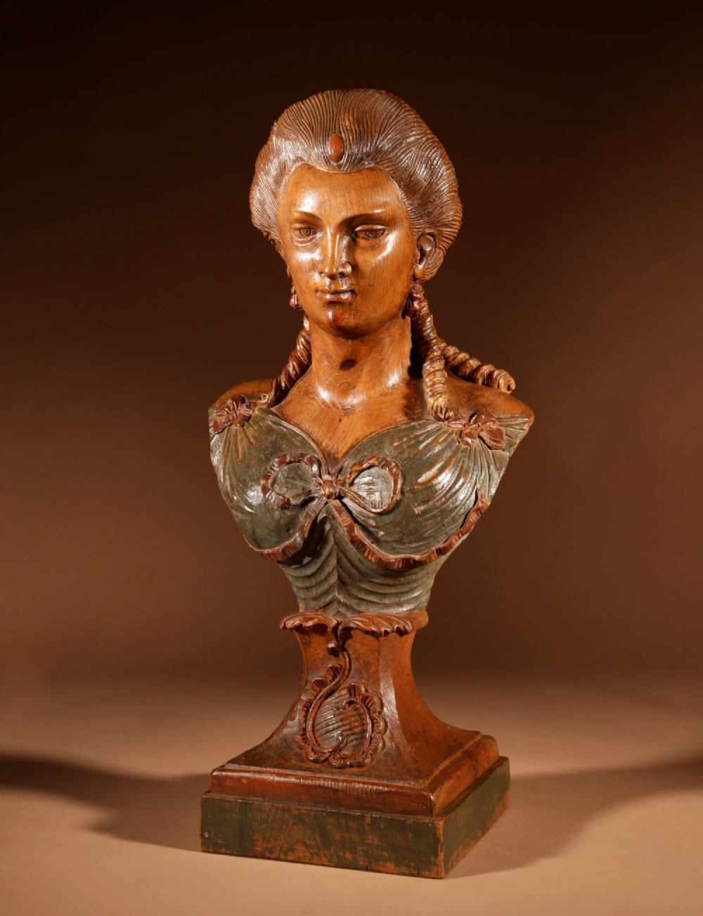 A hand-carved wooden bust of a female figure featuring swept-back hair, braided locks, a draped bodice with bow detail, resting on an integrated pedestal base.