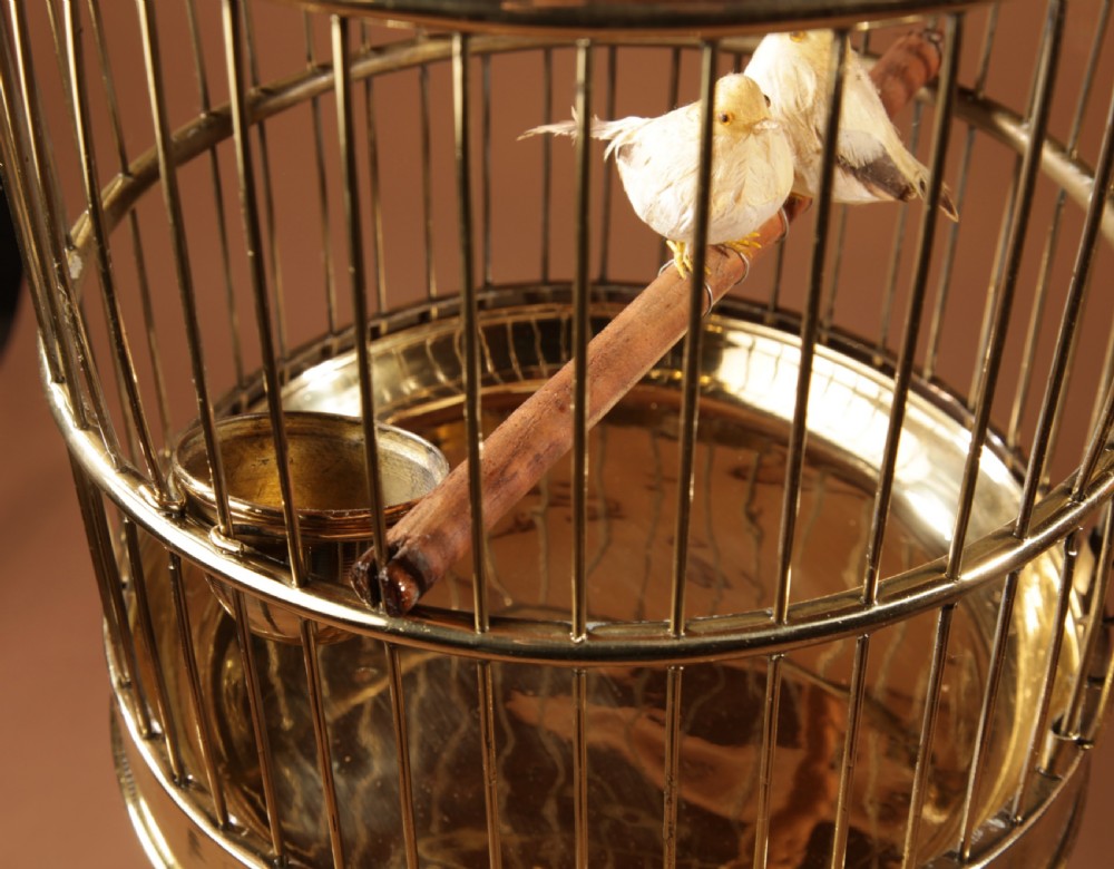 A polished round brass bird cage featuring a domed top, a dark turned wooden finial, and an interior perch with artificial birds. View VI