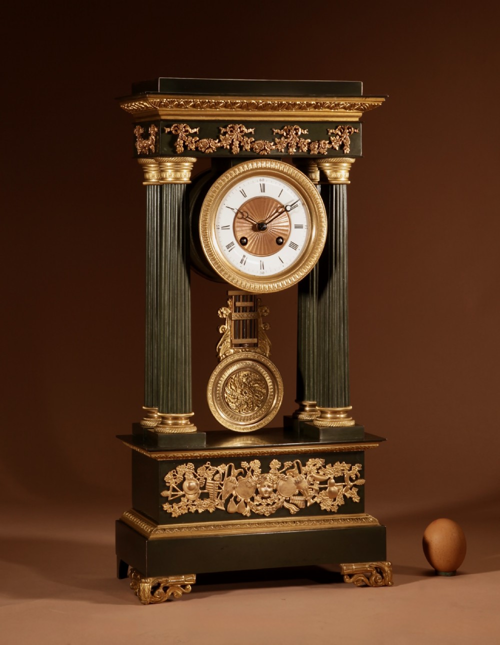 A late nineteenth-century French portico clock featuring dark patinated bronze columns, decorative gilt bronze mounts, a white enamel dial with Roman numerals, and an ornate pendulum.