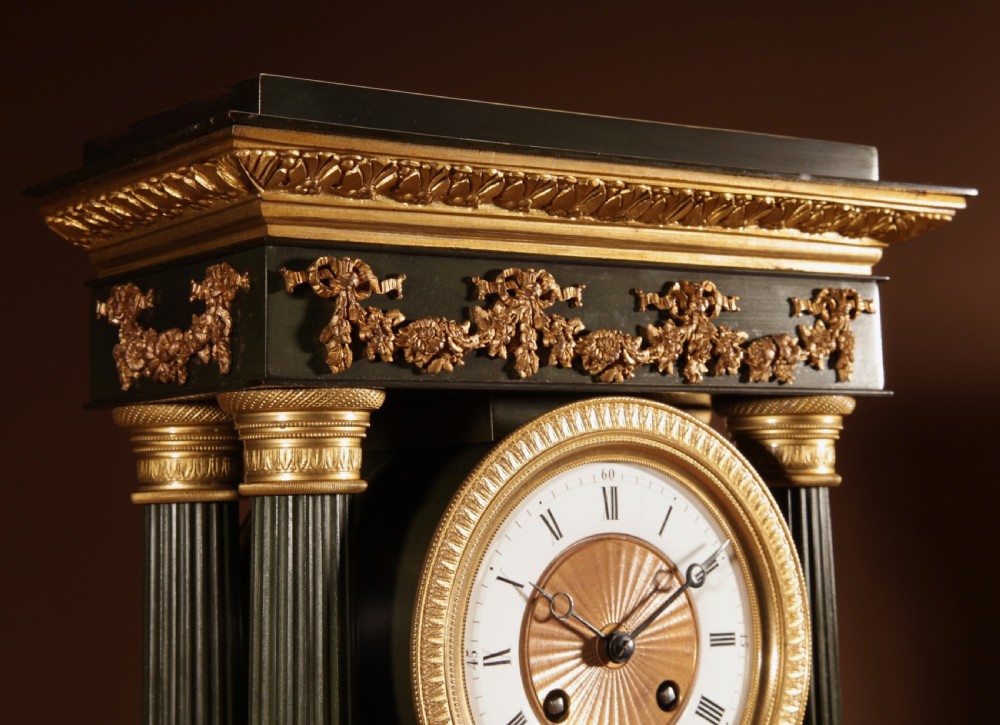 A late nineteenth-century French portico clock featuring dark patinated bronze columns, decorative gilt bronze mounts, a white enamel dial with Roman numerals, and an ornate pendulum. View V