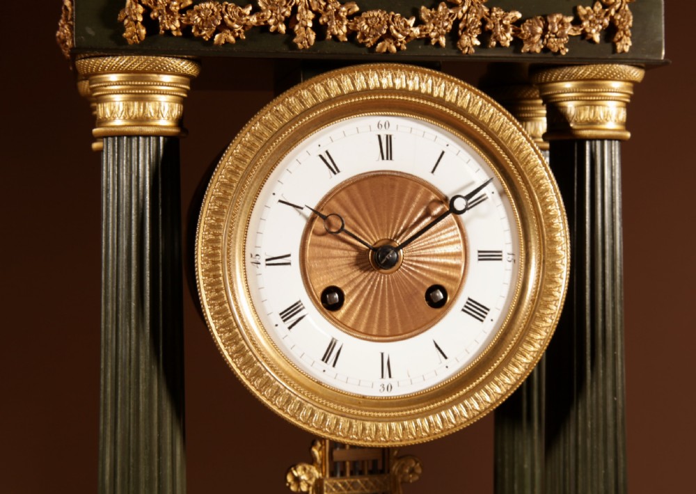 A late nineteenth-century French portico clock featuring dark patinated bronze columns, decorative gilt bronze mounts, a white enamel dial with Roman numerals, and an ornate pendulum. View VI