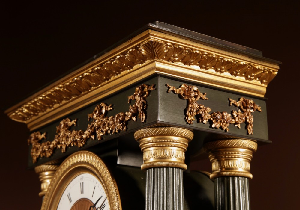 A late nineteenth-century French portico clock featuring dark patinated bronze columns, decorative gilt bronze mounts, a white enamel dial with Roman numerals, and an ornate pendulum. View XII