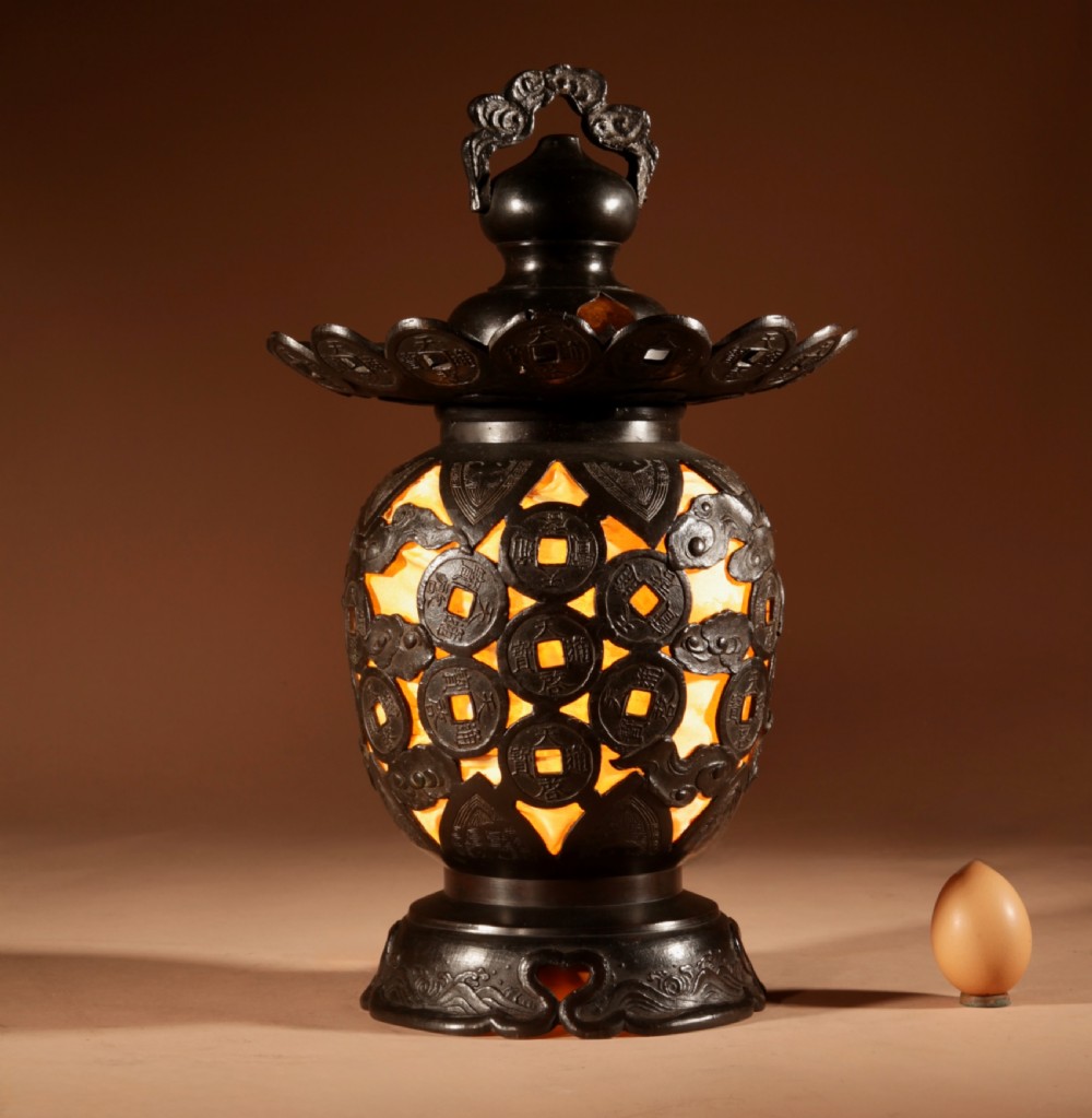 A dark bronze Japanese table lantern featuring reticulated coin and cloud pattern cut-outs, an illuminated amber interior, and a scalloped crest with a top handle.