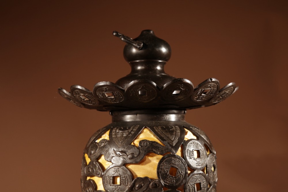 A dark bronze Japanese table lantern featuring reticulated coin and cloud pattern cut-outs, an illuminated amber interior, and a scalloped crest with a top handle. View V