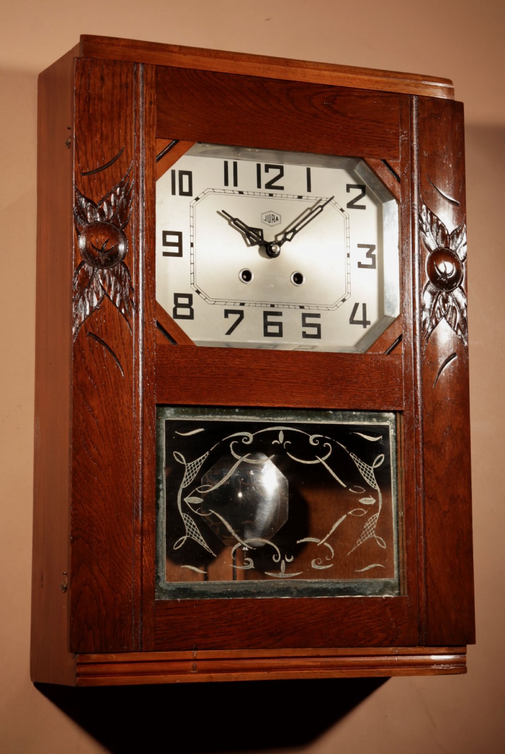 A rectangular Art Deco French oak wall clock featuring stylised side carvings, a silvered octagonal-framed dial face, and an etched lower glass window revealing a metal pendulum. View III