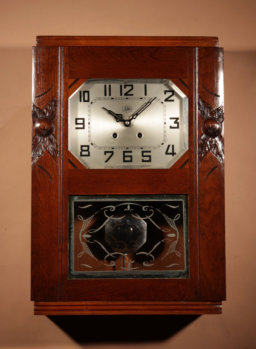A rectangular Art Deco French oak wall clock featuring stylised side carvings, a silvered octagonal-framed dial face, and an etched lower glass window revealing a metal pendulum. View IV