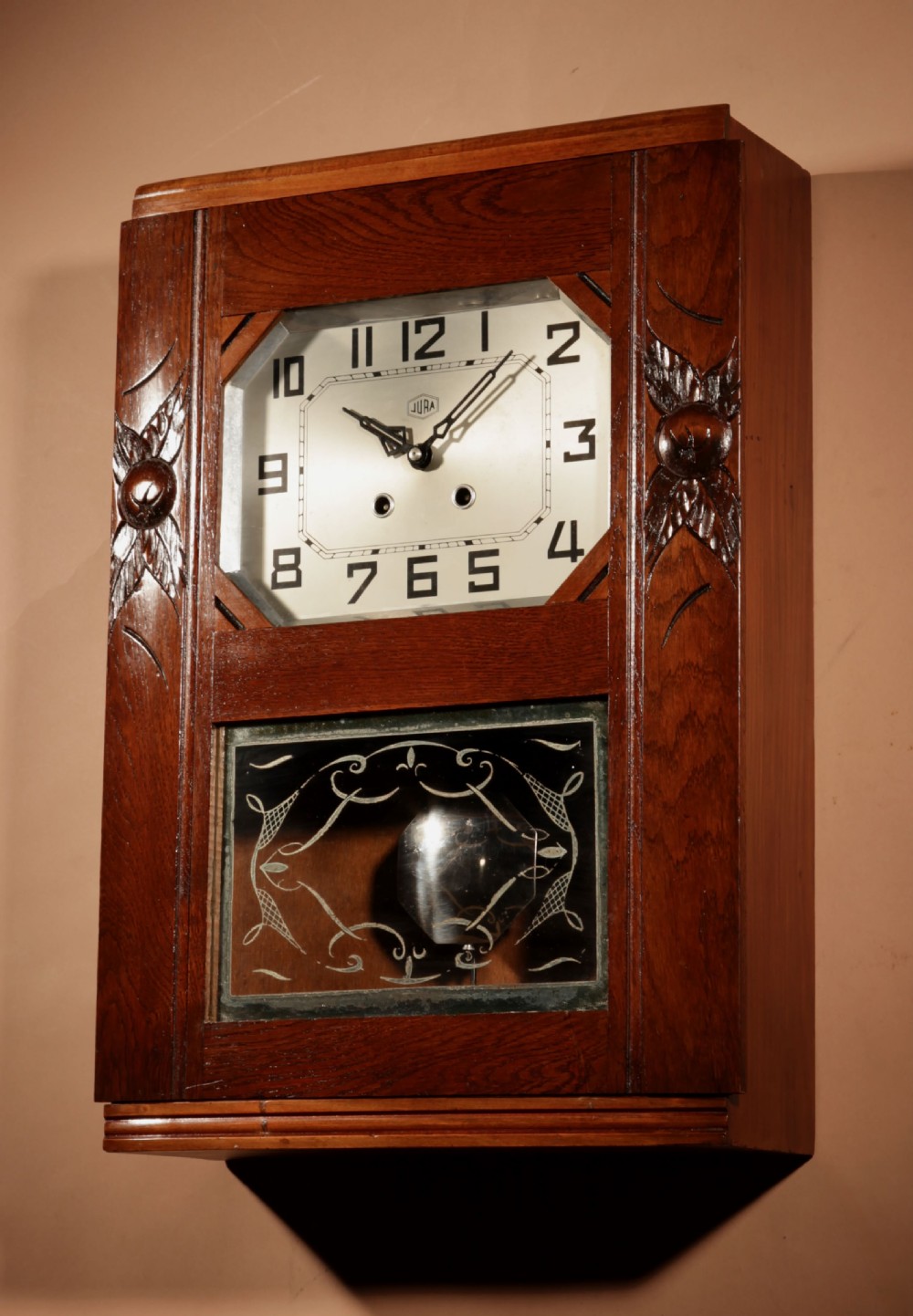 A rectangular Art Deco French oak wall clock featuring stylised side carvings, a silvered octagonal-framed dial face, and an etched lower glass window revealing a metal pendulum. View V