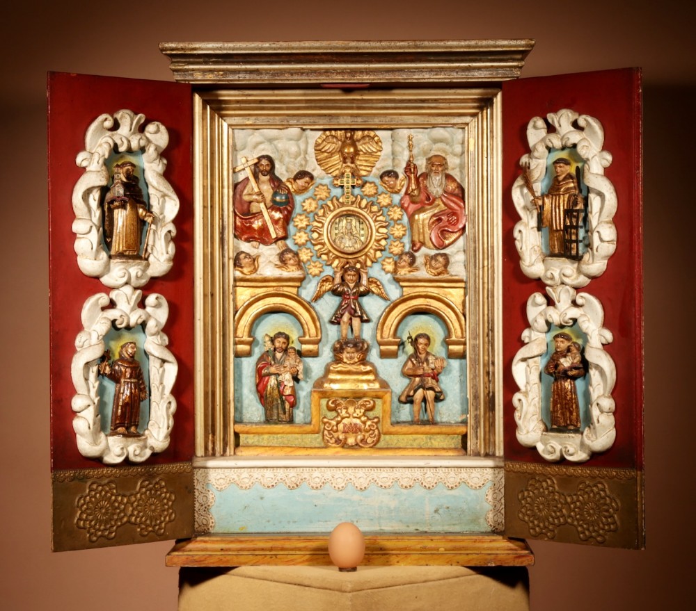 A polychrome wooden Spanish home altar with open red doors featuring carved saint figurines surrounding a gilded central shrine.