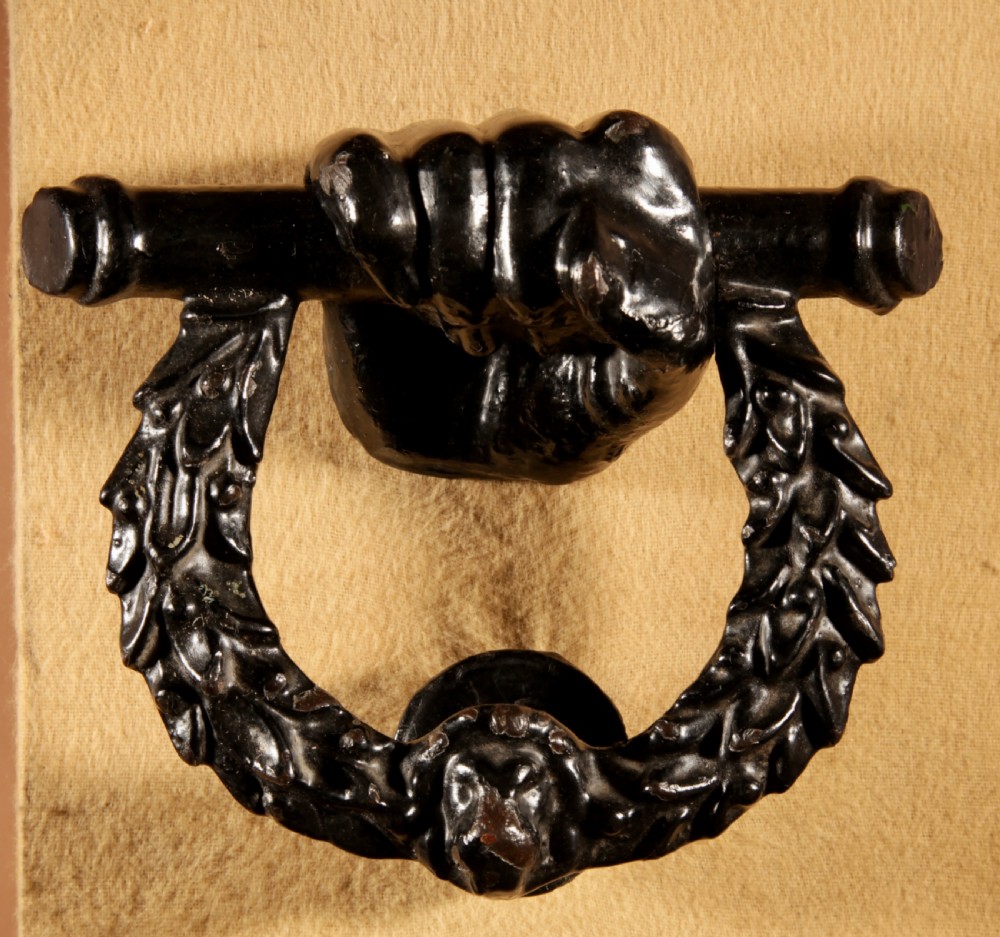 A black cast iron door knocker featuring a clenched hand holding a horizontal rod above a laurel wreath ring with a central lion head mask.