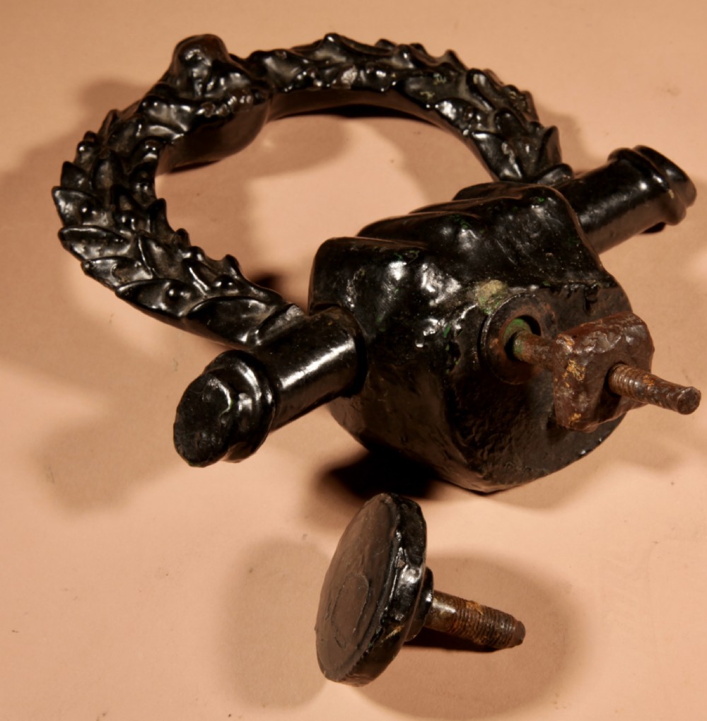 A black cast iron door knocker featuring a clenched hand holding a horizontal rod above a laurel wreath ring with a central lion head mask. View III