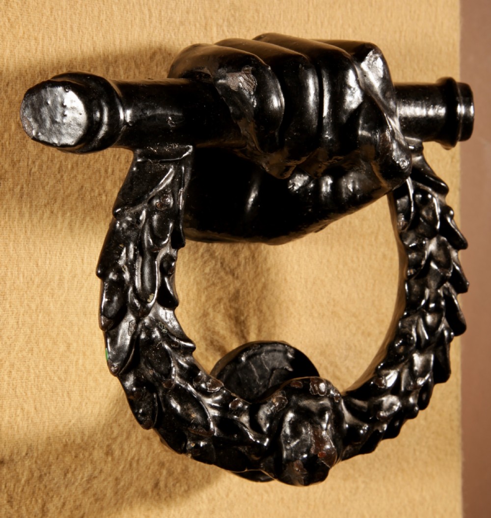 A black cast iron door knocker featuring a clenched hand holding a horizontal rod above a laurel wreath ring with a central lion head mask. View IV