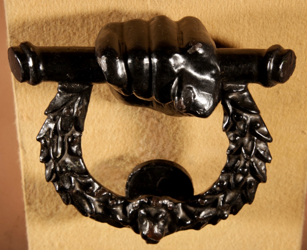 A black cast iron door knocker featuring a clenched hand holding a horizontal rod above a laurel wreath ring with a central lion head mask. View VI