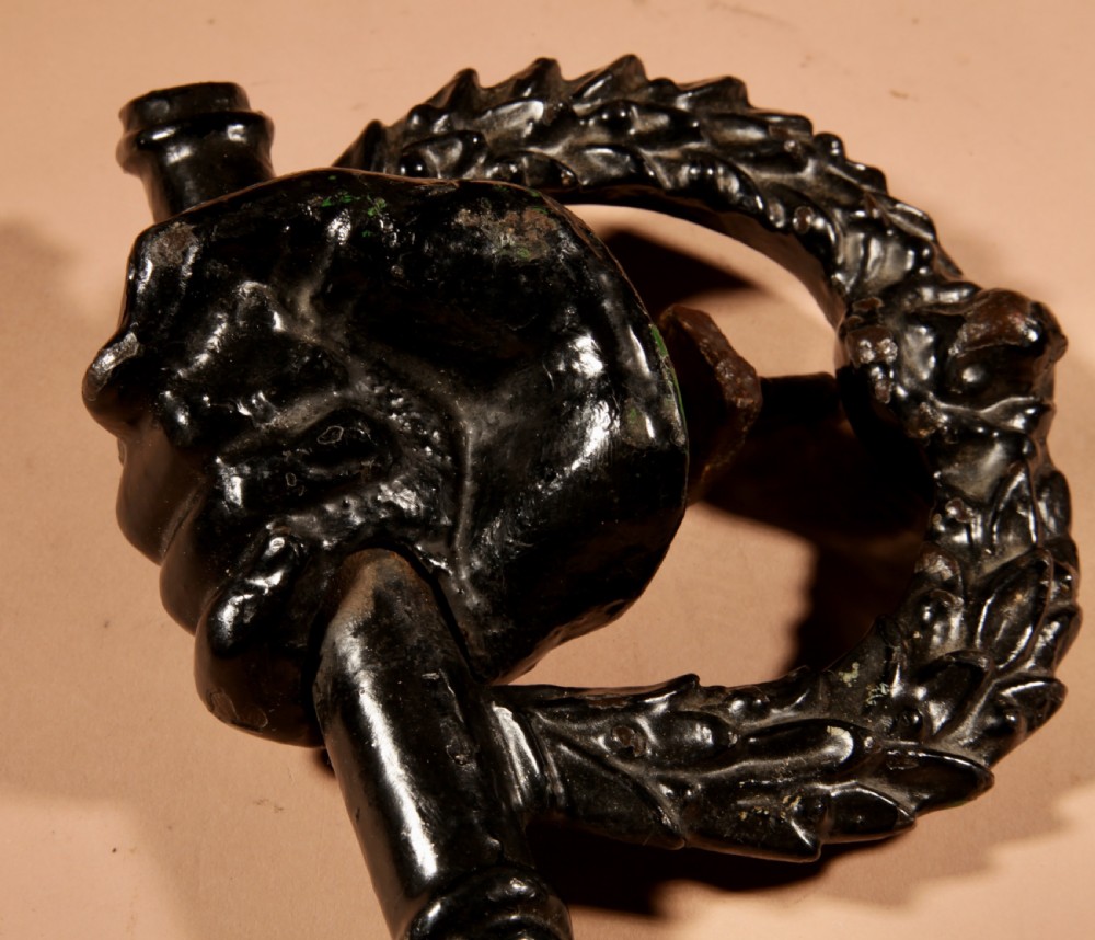 A black cast iron door knocker featuring a clenched hand holding a horizontal rod above a laurel wreath ring with a central lion head mask. View VIII