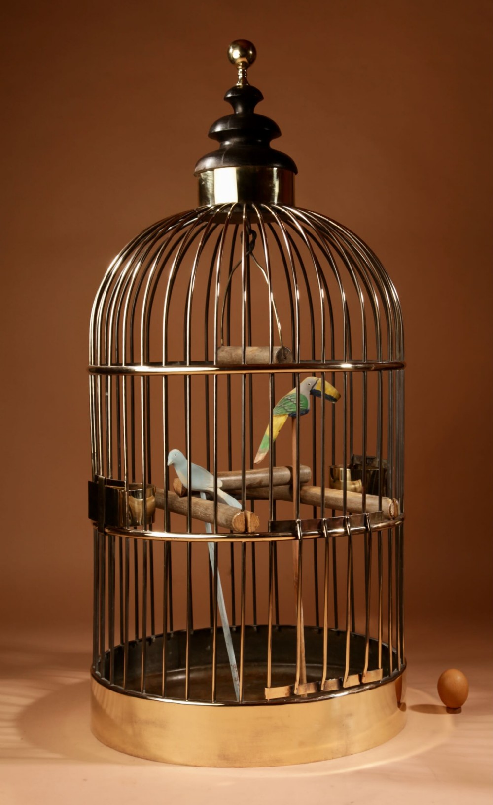A large, cylindrical antique brass bird cage featuring a domed wired top, a turned wooden finial, interior perches, and carved decorative wooden birds.
