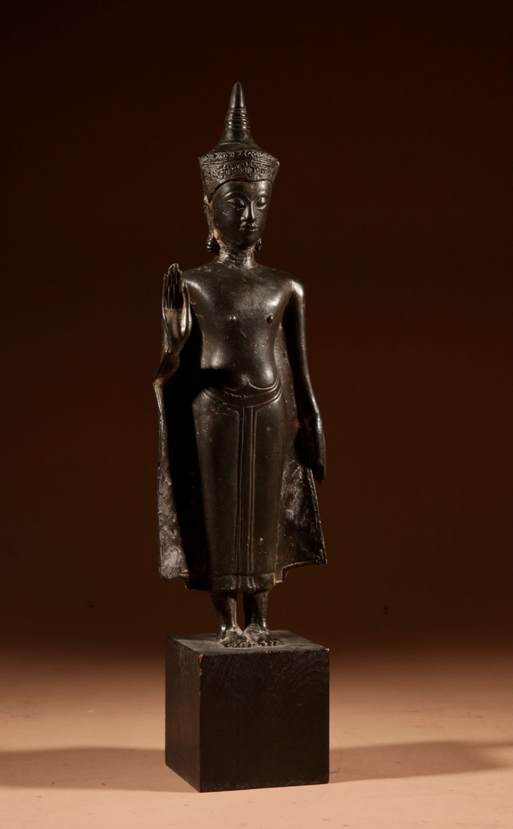 A dark bronze standing crowned Buddha figure depicted in the Abhaya mudra pose mounted on a dark square base.
