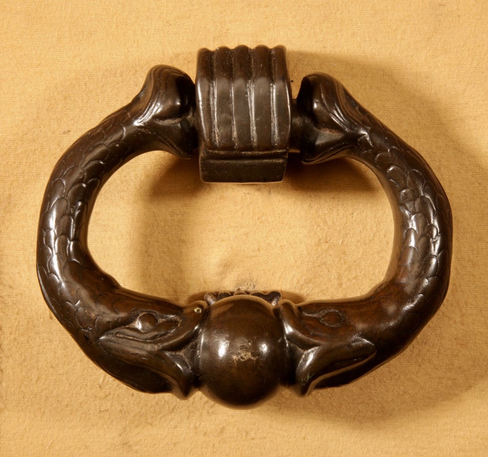 A cast dark bronze door knocker featuring stylized dolphin or serpent figures joining at a central sphere with a ribbed hinge block.