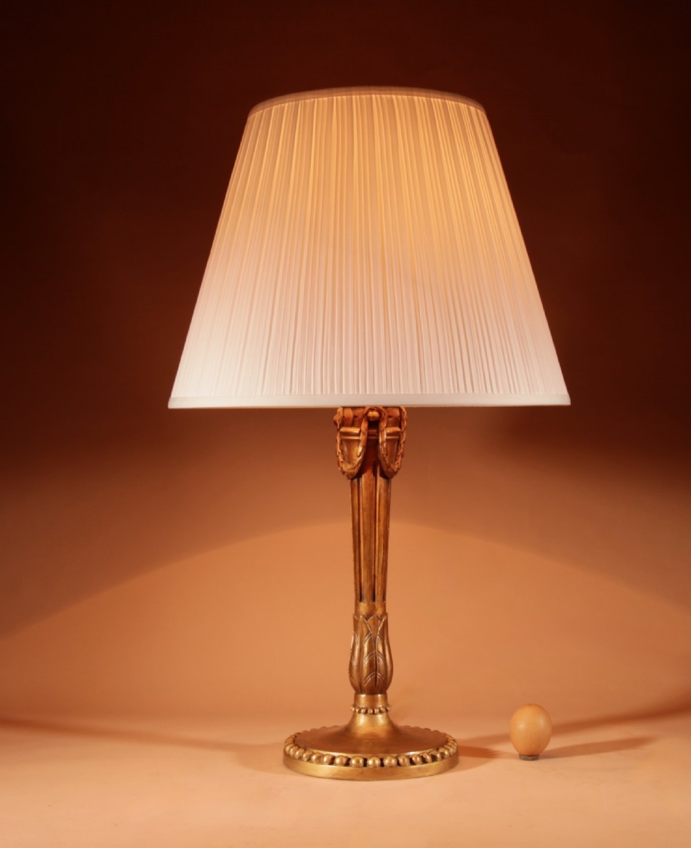 A Louis XVI style carved giltwood table lamp featuring a fluted stem, beaded base, and cream pleated shade.