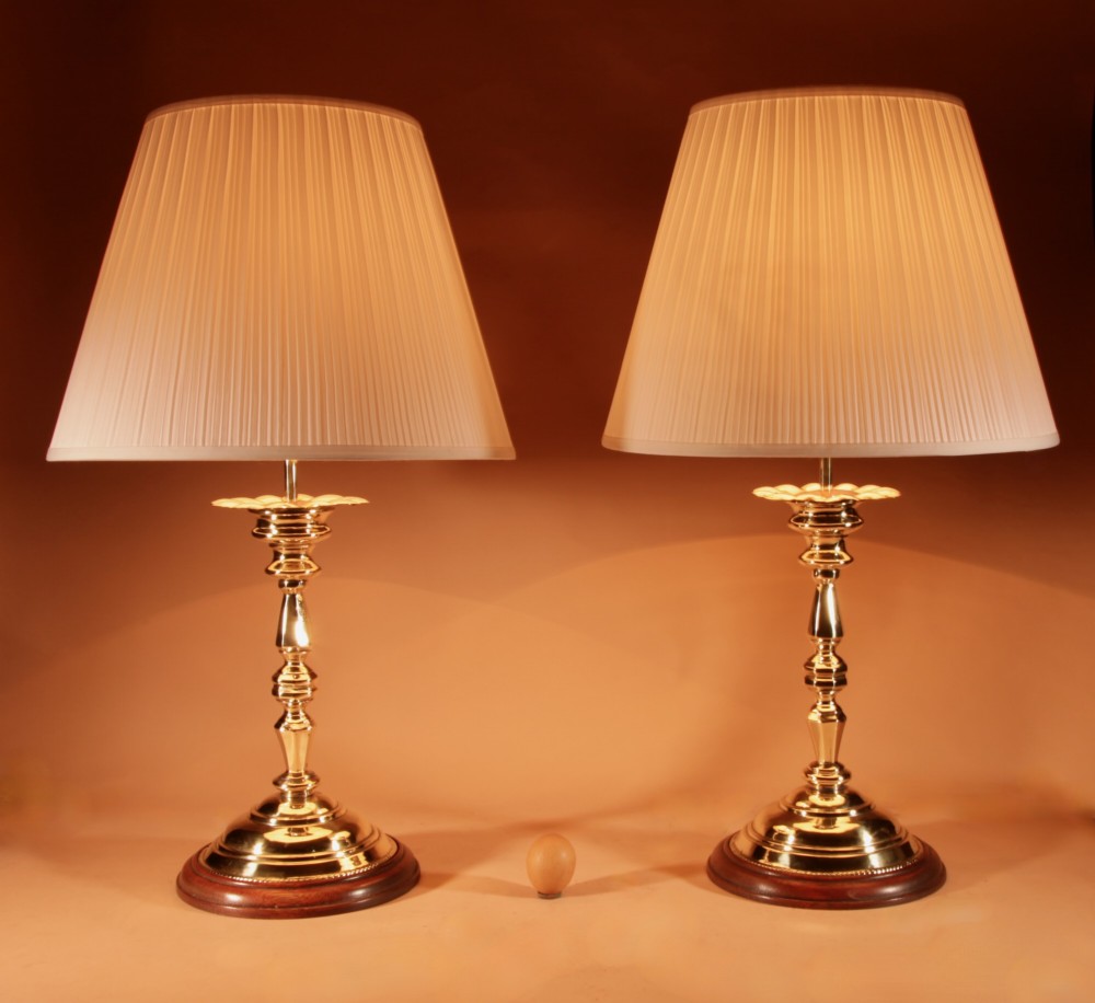 A pair of turned brass candlestick-style table lamps set on circular wooden bases with cream pleated shades.