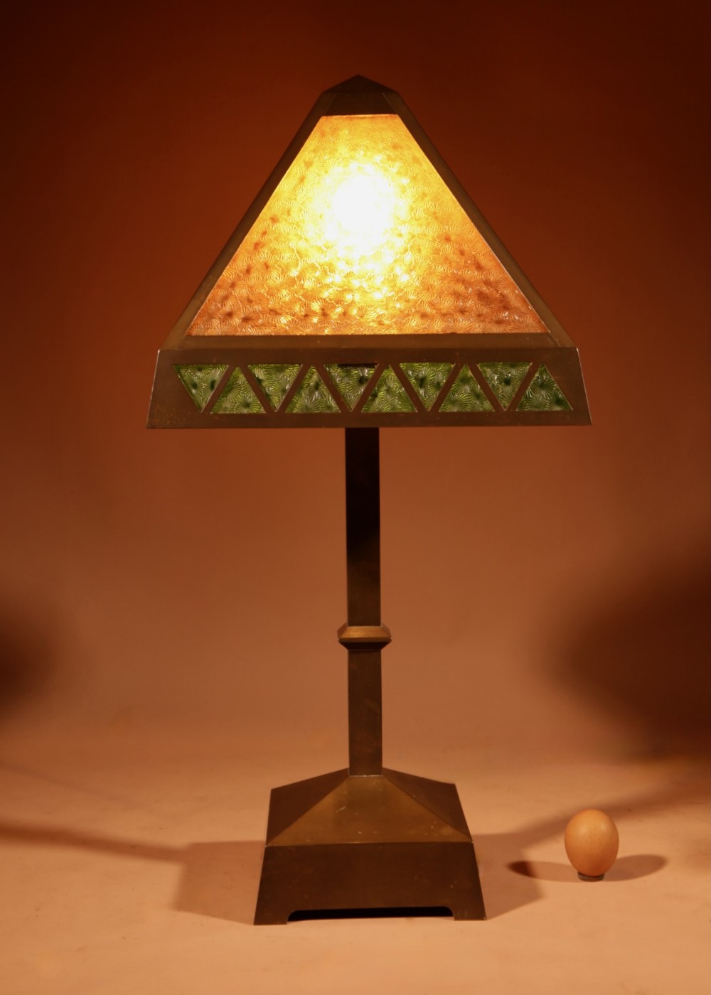 An antique patinated brass table lamp featuring a pyramidal shade with amber textured glass and a decorative band of green triangular glass panels.