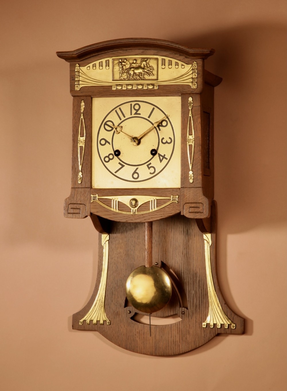 An Art Nouveau light oak wall clock featuring brass mounts, an embossed chariot motif top panel, and a visible pendulum.